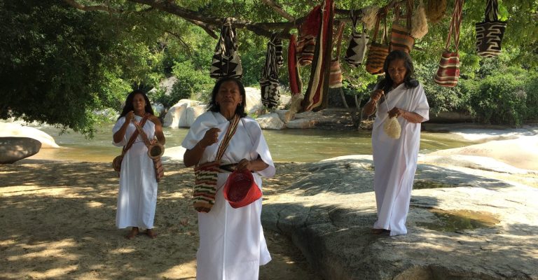 Trois femmes en vêtements traditionnels se tiennent près d'une rivière, entourées de nature.